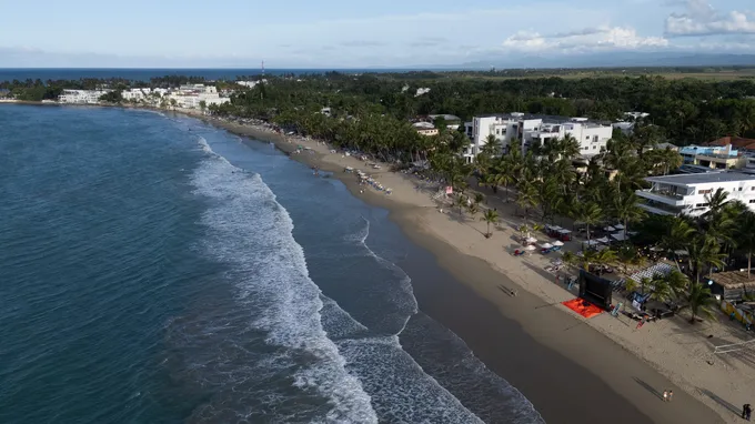 Aerial view of Cabarete beach, with surf breaking along the curved shore and low white buildings behind the palm trees.